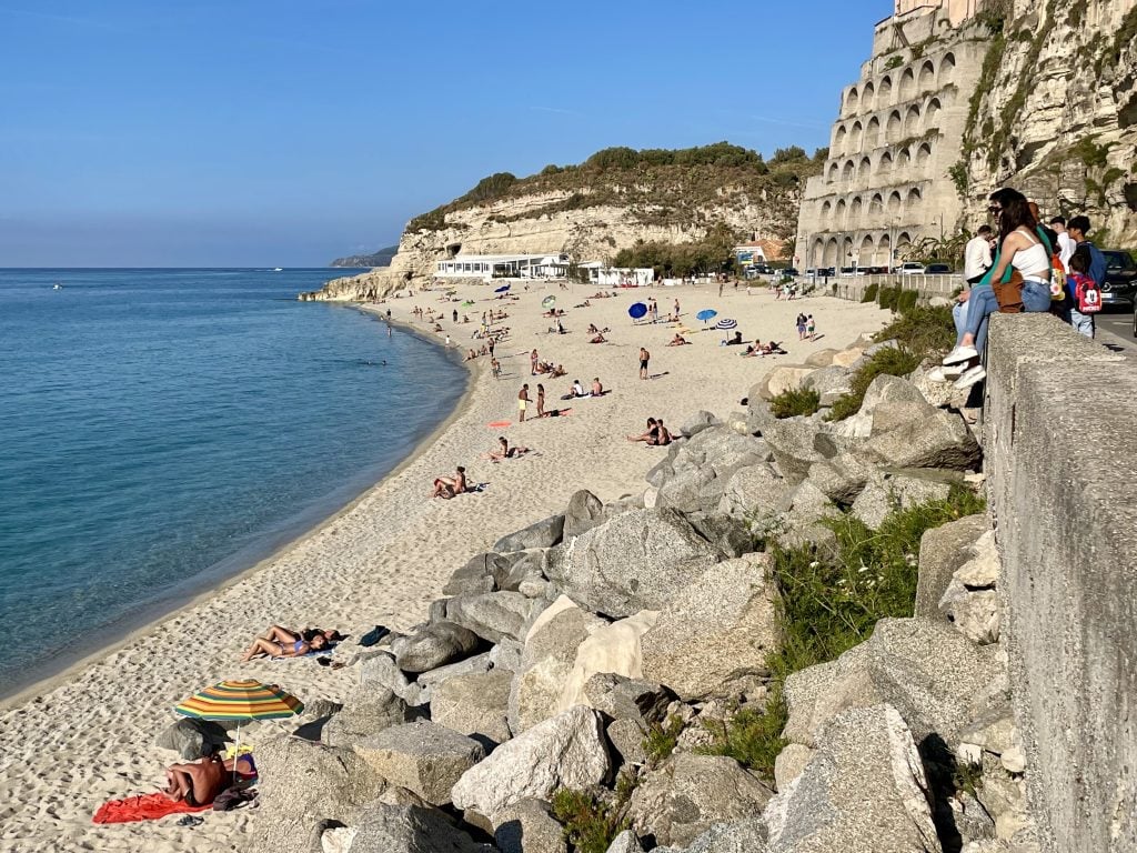 A view of Tropea's long, sandy beach, lots of people relaxing on the sand, and a girl sitting on a wall watching the people on the beach.