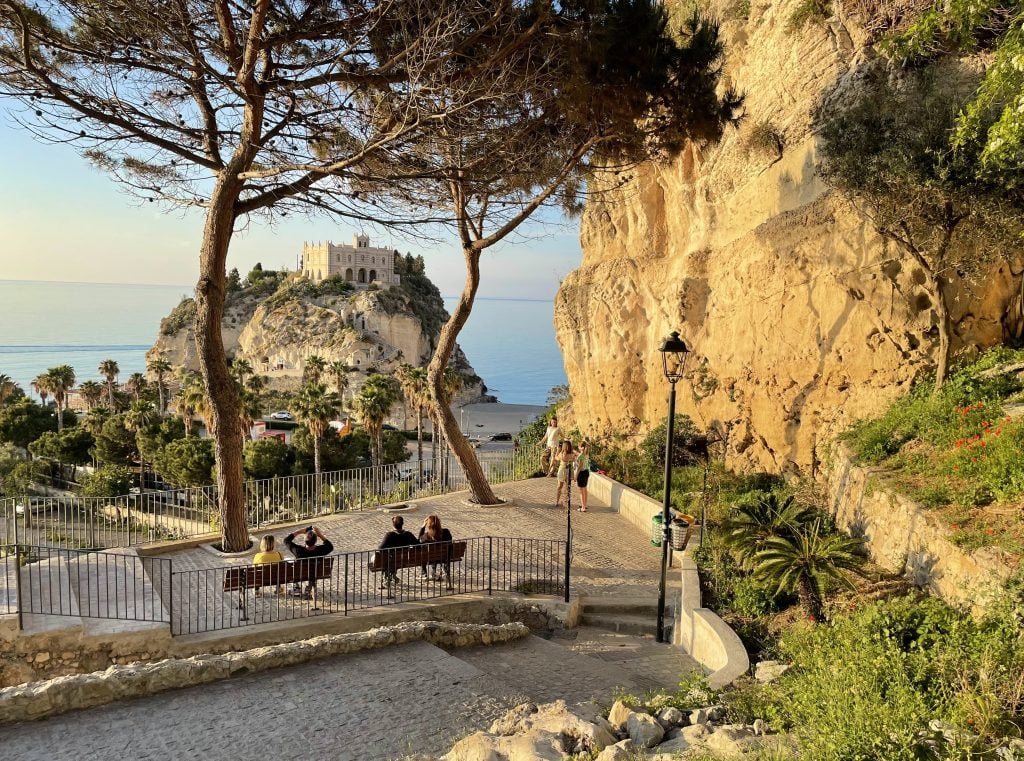 A view of the sanctuary along the water in the background. In the foreground, four people sitting on two benches on a tiny mini-piazza enjoying the view. A woman poses for photos as her friends take shots.