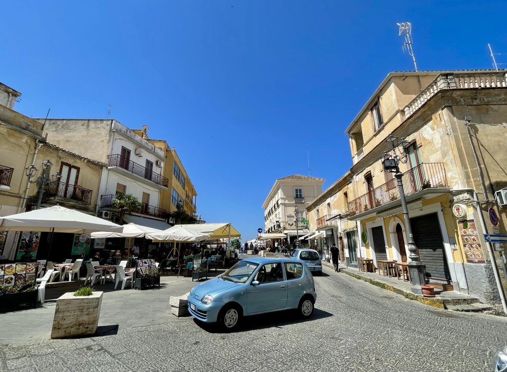 A piazza in Pizzo, Italy, lots of sidewalk cafes around. In the center is a small baby blue car.