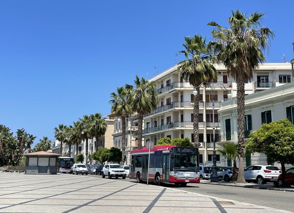 A street lined with palm trees and tall white buildings with balconies in Reggio Calabria. A red bus barrels down the street.