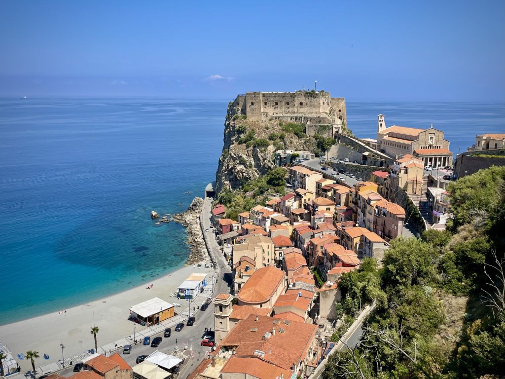 The town of Scilla, a dark gray rock castle sitting atop a rocky cliff, and homes with orange roofs along the hillside. There is a beach with clear blue water to the left of the castle and homes.