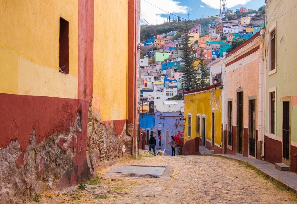 A small hilly cobblestone street giving way to hills in the background with even more colorful buildings.