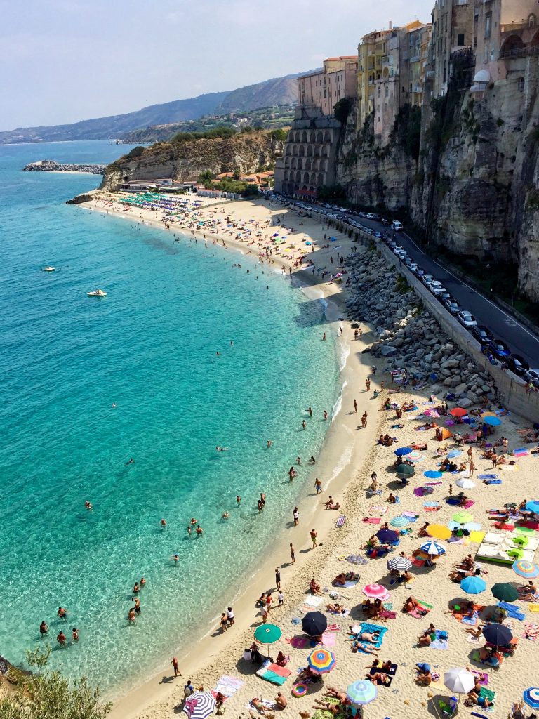 A beach in Tropea, Italy with neon turquoise clear water. Hundreds of people on the beach with bright umbrellas and towels. The beach is next to a tall stone cliff topped with buildings.