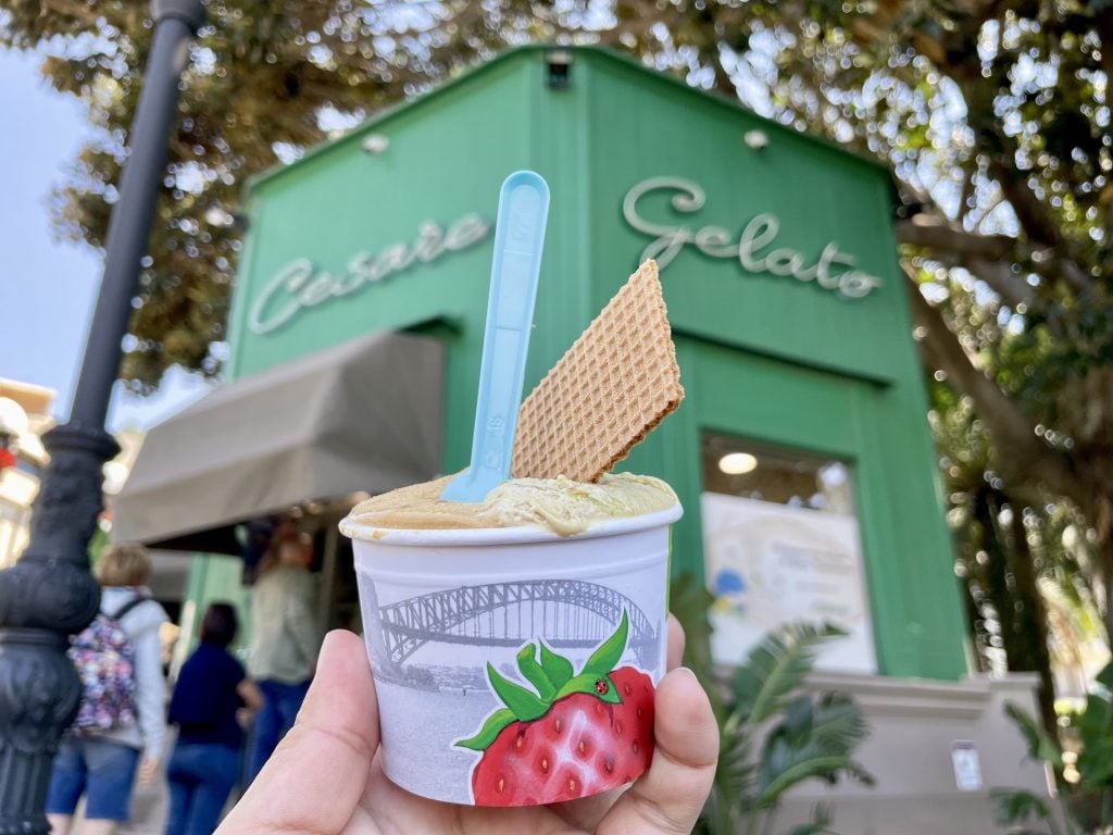 A hand holding a cup of pistachio gelato with a spoon sticking out of it in front of a green kiosk-type building reading "Cesare Gelato."