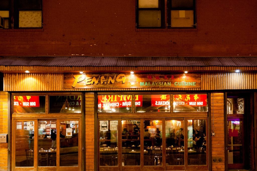 Penang Malaysian Restaurant in Boston's Chinatown, lit up in warm light after dark, windows reflecting Chinese restaurants across the street.