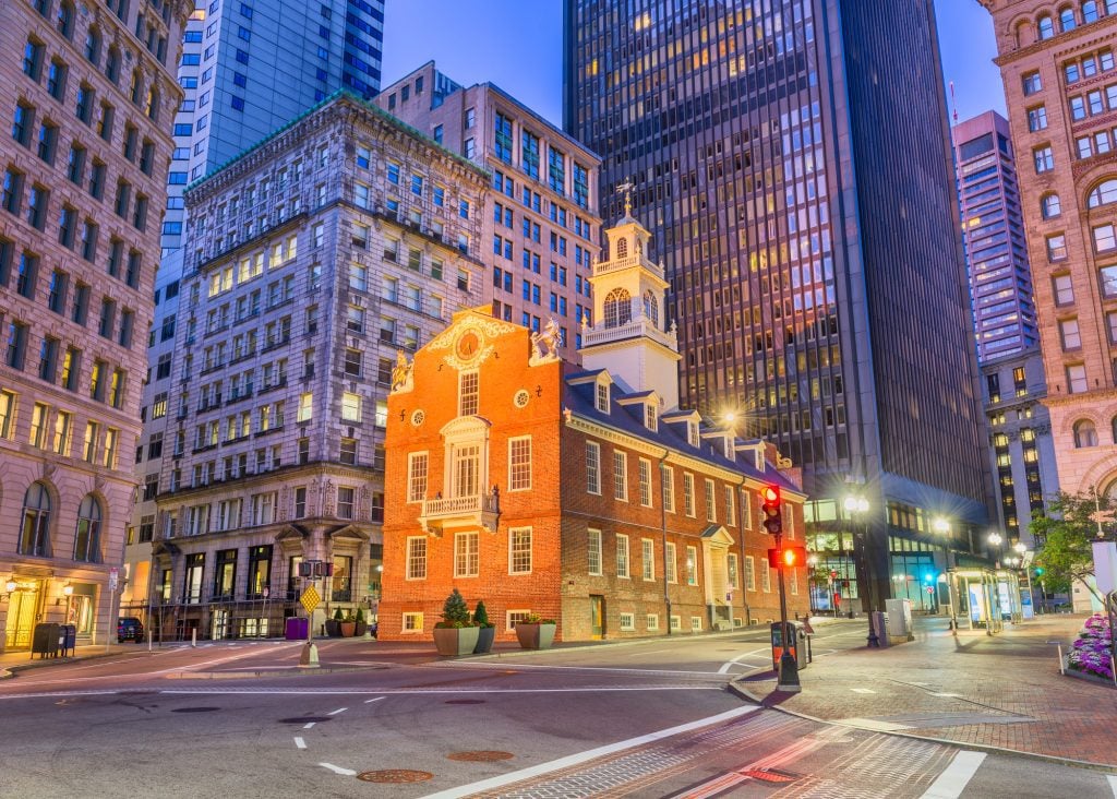 A brightly lit up 18th century brick building with a white bell tower, surrounded by modern skyscrapers at twilight.