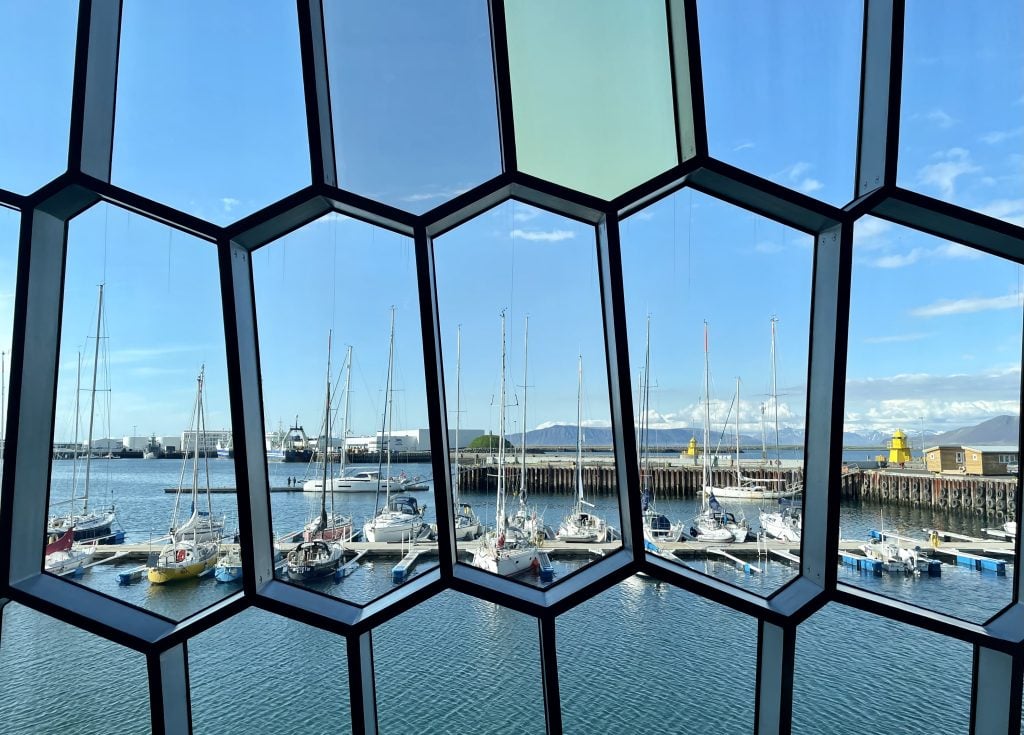 Several sailboats docked in a calm harbor, as seen from inside Harpa. You are looking through several windows shaped like elongated hexagons fitted together.