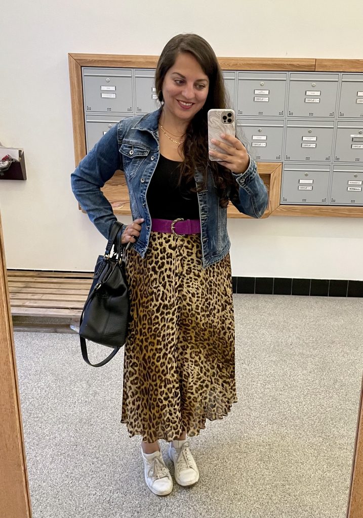 Kate takes a smiling mirror selfie wearing a blue denim jacket, black tank top, bright magenta suede belt with a gold buckle, midi-length pleated leopard skirt, white sneakers, and carrying a black leather bag.