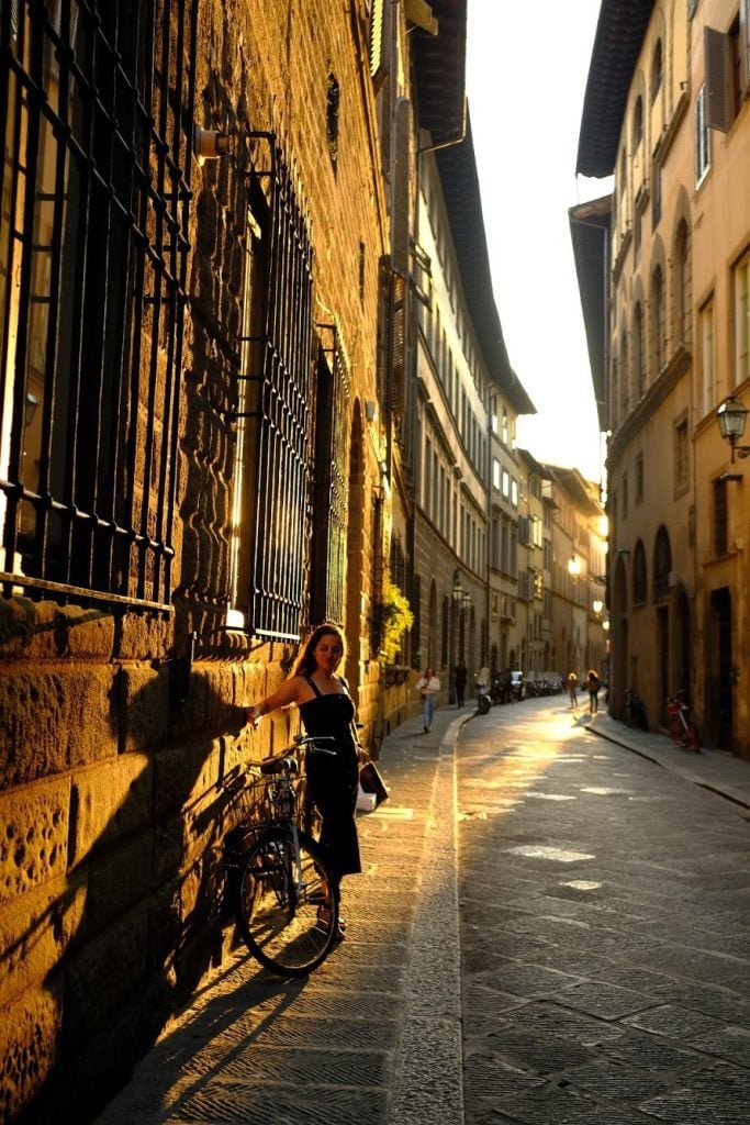 Kate standing on the street in Florence, wearing a black dress and standing beside a bicycle, the late afternoon light throwing shadows over her.