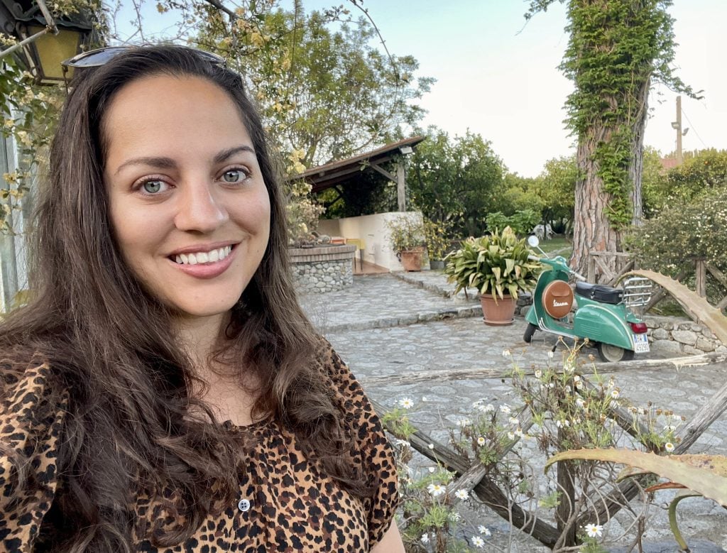 Kate taking a smiling selfie in a leopard-print shirt, behind her is a rural guesthouse with gardens and a turquoise Vespa.