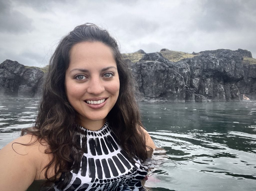 Kate smiles in the gray misty water of the Sky Lagoon. She wears a black and white patterned bathing suit top. Behind her you see gray rocks and a stormy gray sky.