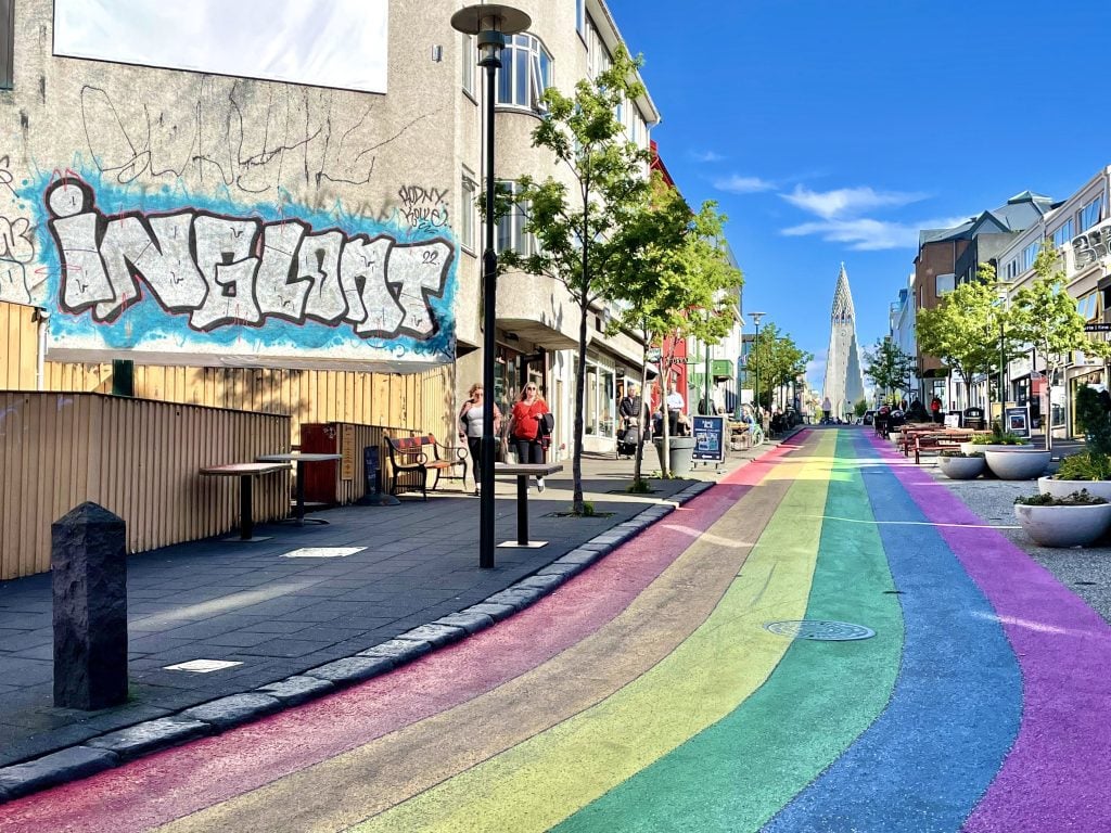 Another view of the rainbow-painted street leading up to Hallgrimskirkja Church. Some boutiques on each side of the street and some graffiti.