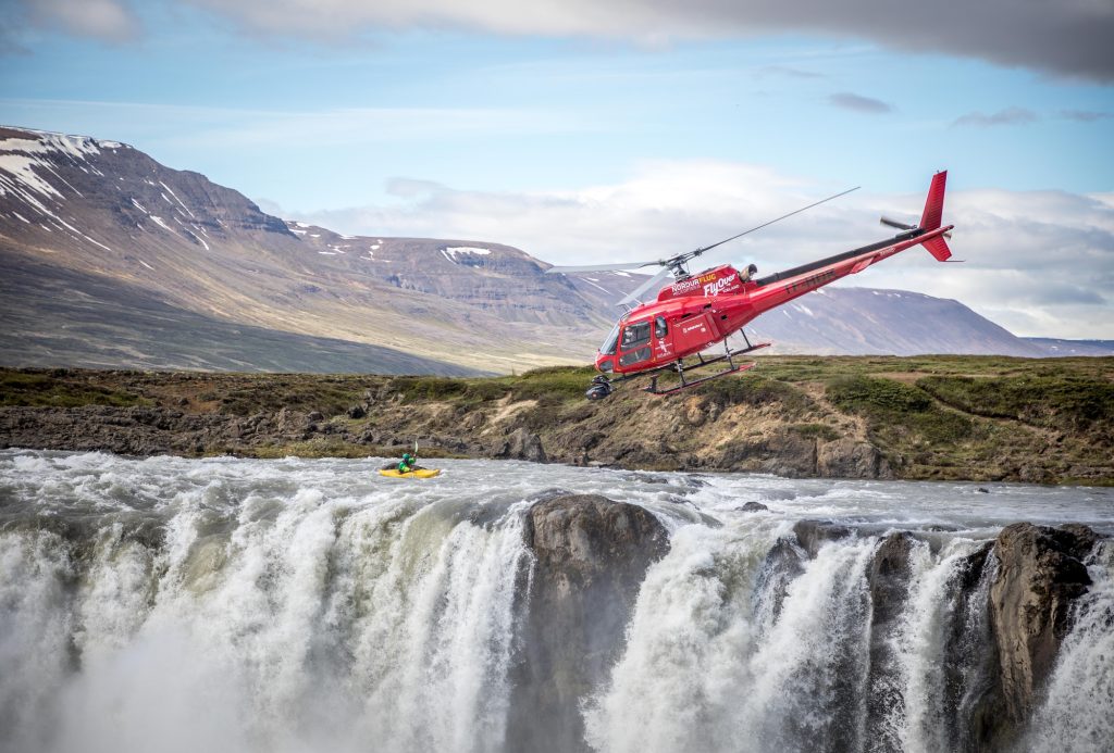 A red helicopter reading "FlyOver" filming a kayaker in a yellow kayak about to go over a huge waterfall.