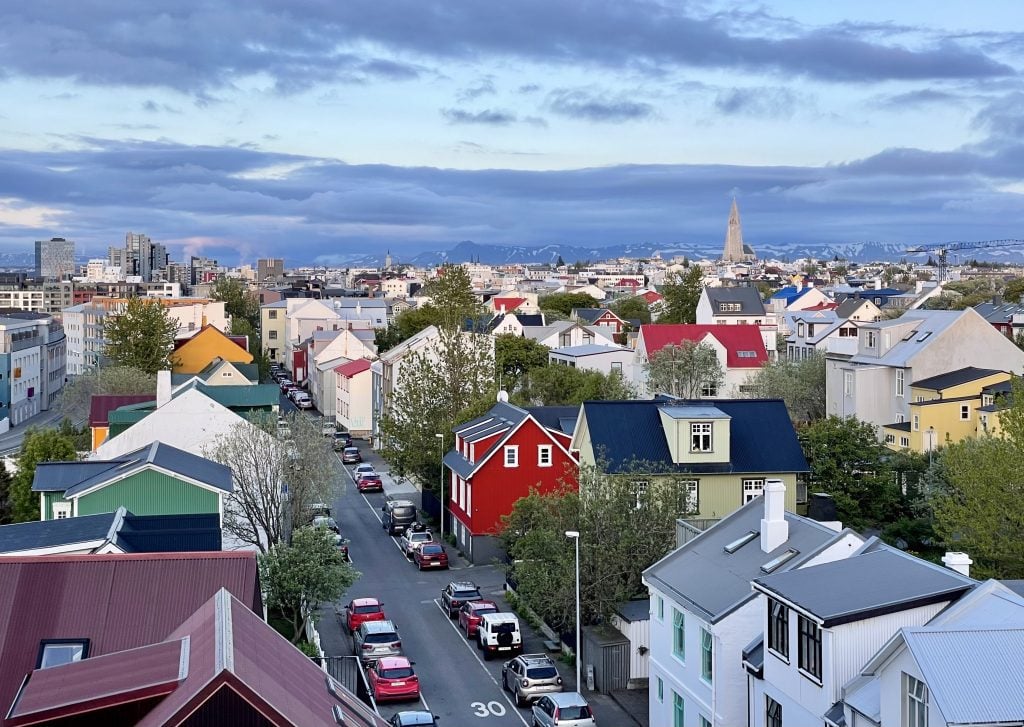 A skyline view of Reykjavik with brightly colored square houses. You see the pointy church on one side and mountains way in the background. The sky is pale blue with dark blue streaky clouds.