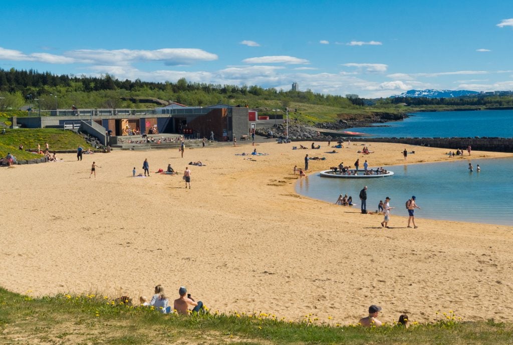 A large golden sandy beach on a sunny day. In the water are a few people; in the water is a large ring filled with hot water with people inside it. On shore is a gray building with a long rectangular hot tub in front, several people inside it.