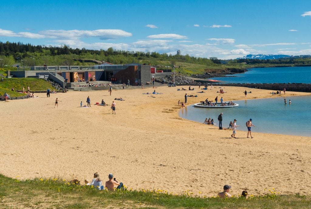 A large golden sandy beach on a sunny day. In the water are a few people; in the water is a large ring filled with hot water with people inside it. On shore is a gray building with a long rectangular hot tub in front, several people inside it.