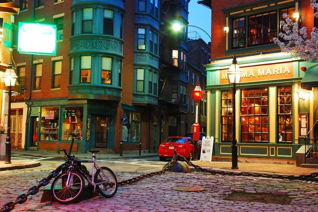 Cobblestone streets, brick buildings, and green rounded balconies, next to Mamma Maria restaurant, in Boston's North End at night.