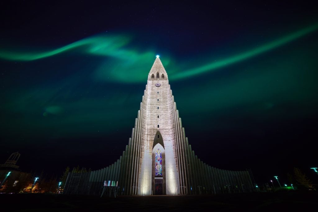 Hallgrimskirkja church in Reykjavik, pointy and gray, and dark green shapes dance behind it (the Northern Lights).