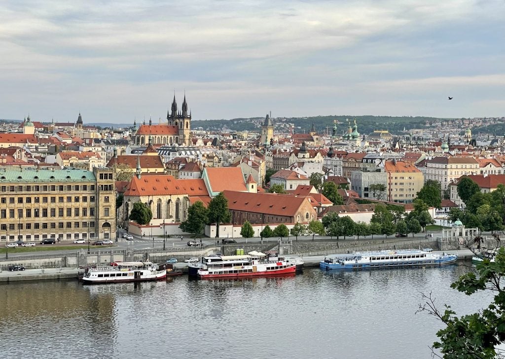 The skyline view of Prague in front of the Vltava River, with lots of white crenellated buildings, orange roofs, and spindly tops of church towers.