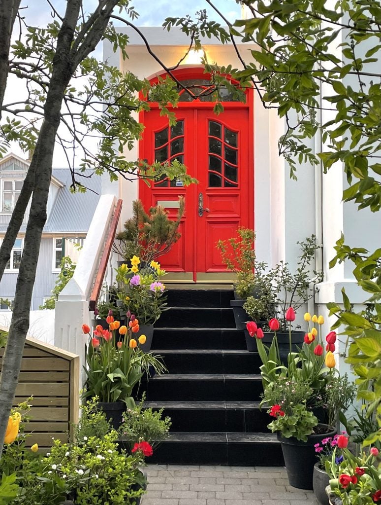 A white building with a curvy red door, surrounded by tulips and other flowers in pots everywhere.
