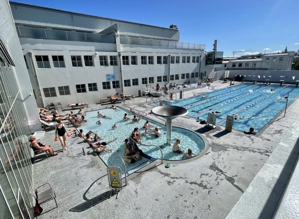 An outdoor complex with a swimming pool where people are swimming laps, a long rectangular-shaped hot tub, and a small shallow pool with kids and adults lying down in it.