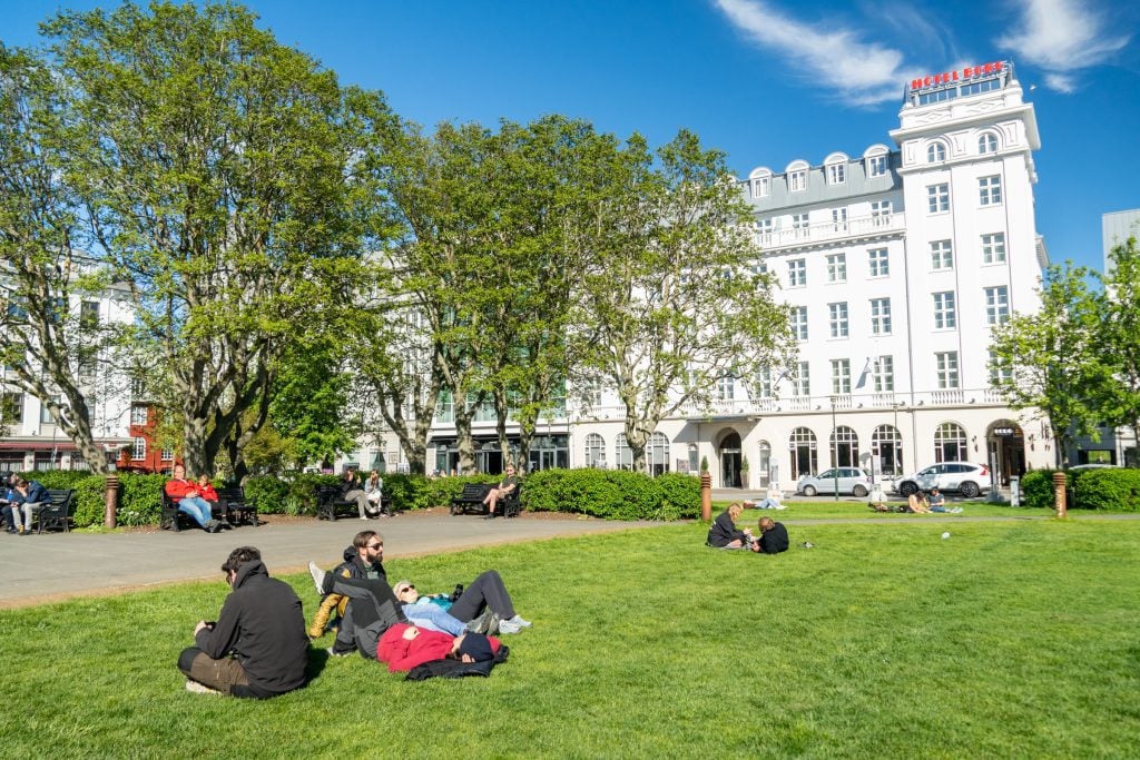 People sitting on the lawn in front of a tall white building in Reykjavik, enjoying the summer sun.