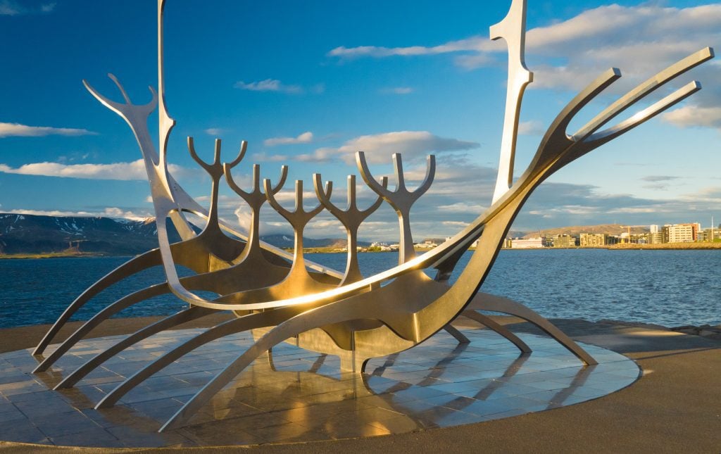 A metal sculpture shaped like a Viking long boat standing next to the shore. It's glinting in the late night sunlight.