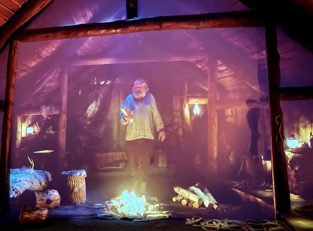 An old man with a big white beard wearing a sweater, holding a log, and standing in front of a fire in a wooden cabin.