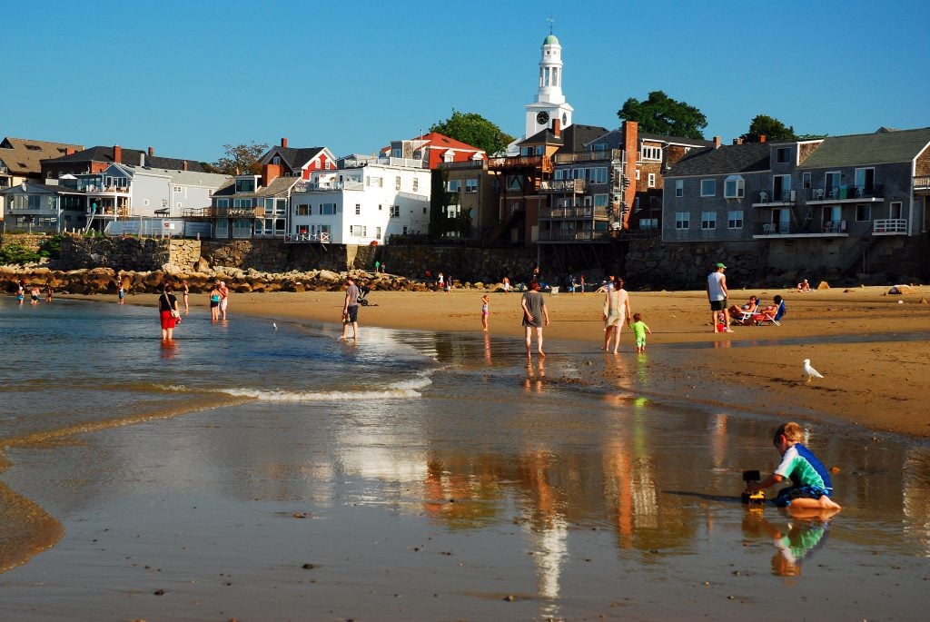 People and kids playing in the shallow waves on a brown sand beach in the afternoon sun, buildings and a church behind them.