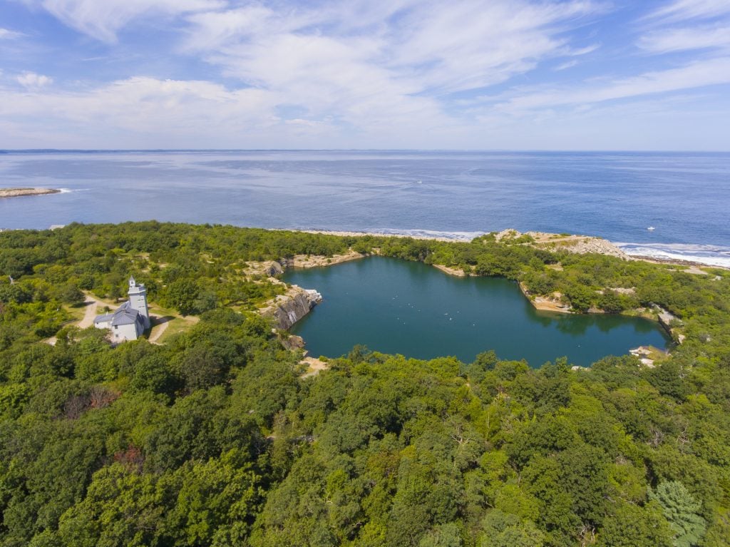 An aerial view of a forested area next to the sea, with a small building and a quarry filled with fresh water.