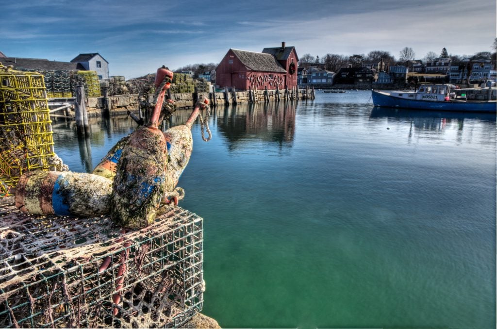 A red cottage on the end of a rocky pier sitting in calm teal water. You see lobster cages and fishing equipment on shore.