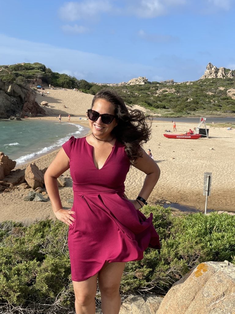 A Guide to La Maddalena, Italy: Fun Things to Do and More! 39 Kate wearing a raspberry colored dress and standing in front of a beach in Sardinia. It's very windy and the dress and her hair are flying.