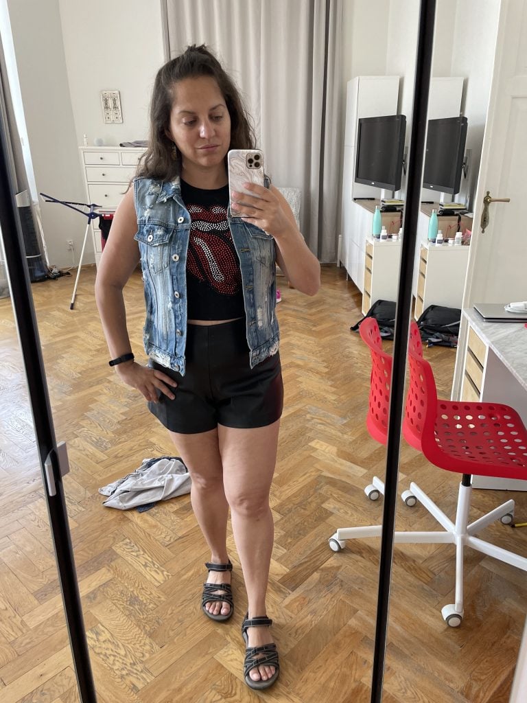 Kate takes a full-length mirror selfie wearing a black crop top with The Rolling Stones giant lips and tongue logo on it, as well as a pale blue distressed denim vest, and short black pleather shorts.