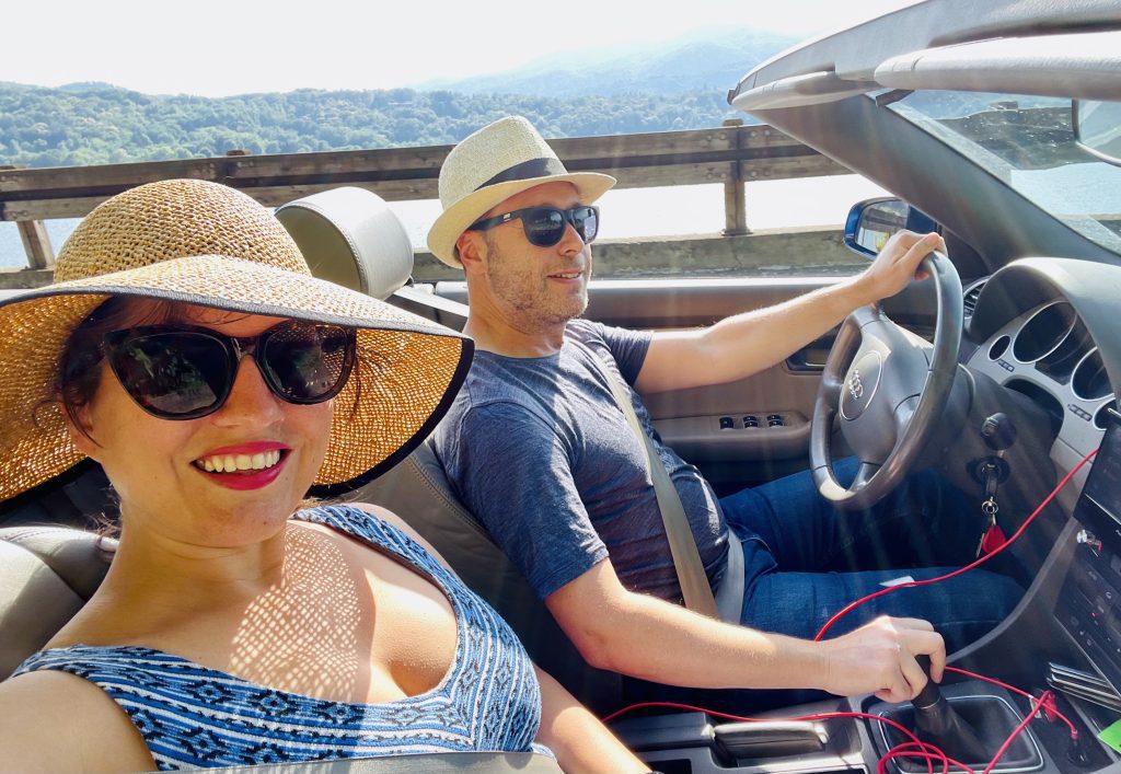 Kate and Charlie wearing sun hats and sunglasses and driving in Charlie's convertible in front of Lake Orta.