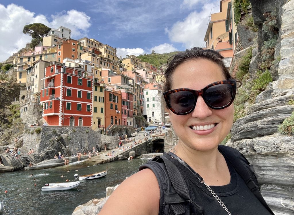 Kate takes a smiling sunglasses selfie in front of narrow colorful buildings in front of a tiny harbor.