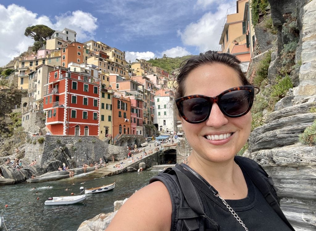 Kate takes a smiling sunglasses selfie in front of narrow colorful buildings in front of a tiny harbor.