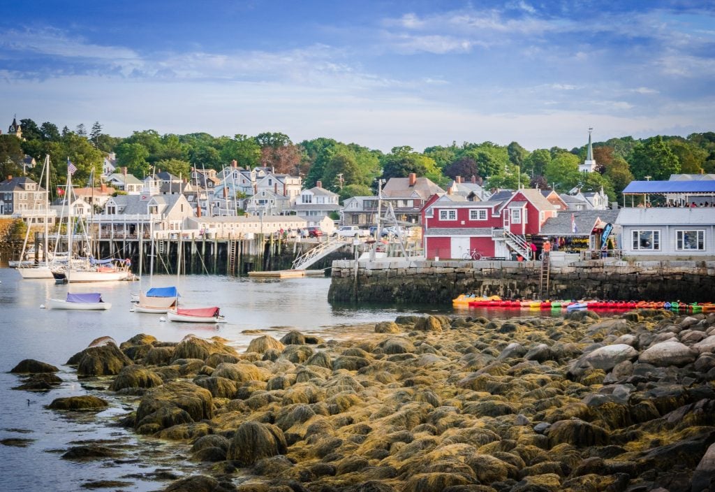 A small seaside town with mostly gray cottages on shore, a rocky coastline, and a few sailboats in the harbor.