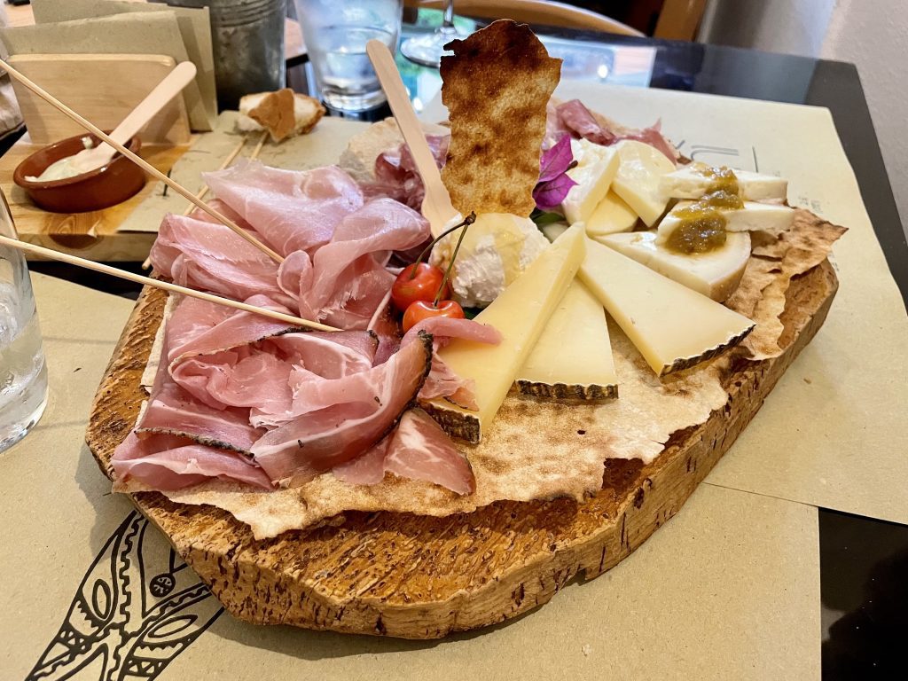 A wooden platter covered with Sardinian meats, cheeses, two cherries, and traditional flatbread.