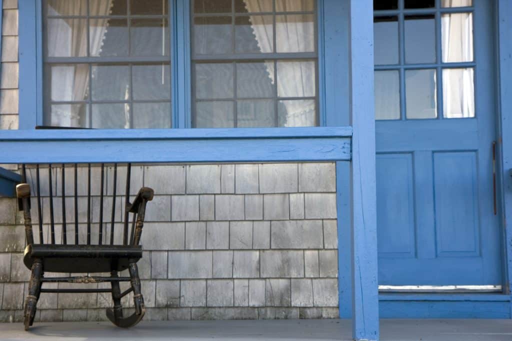 A single wooden rocking chair on the porch of a gray house with bright blue trim.