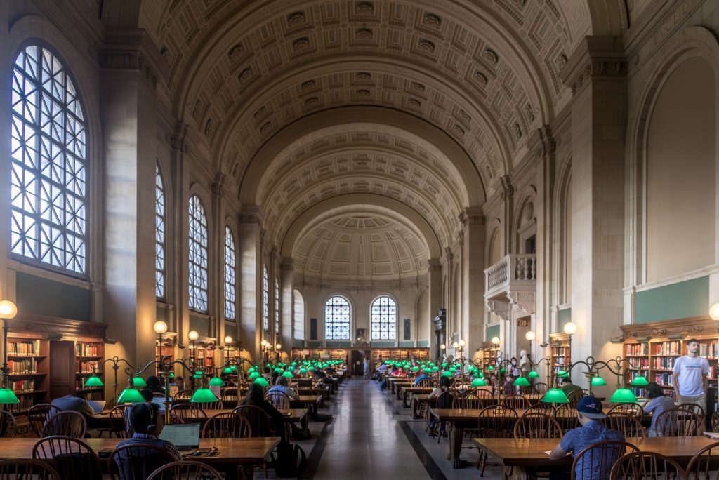 A large, long library room with a curved granite ceiling. Rows of tables with green lamps, people studying at them.