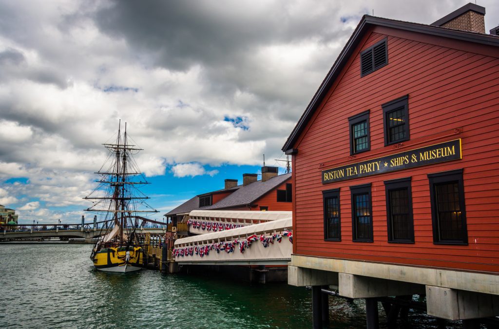 A red house reading Boston Tea Party Ships & Museum next to the water, where a yellow ship is anchored.