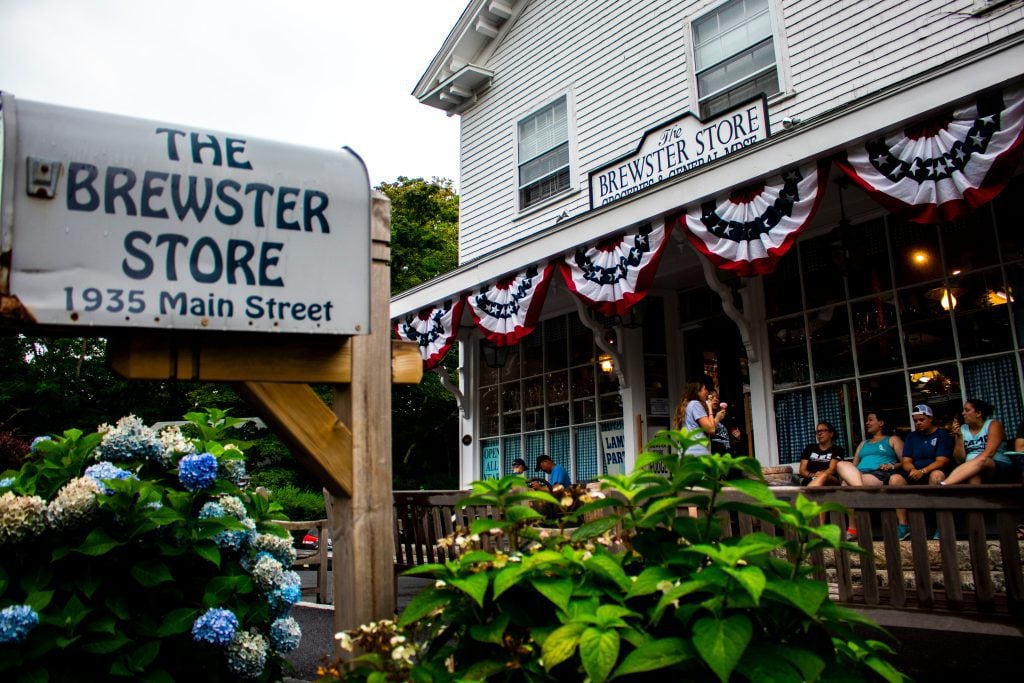 A white store with a sign reading The Brewster Store, people sitting on a bench on its porch, hydrangea bushes in front.
