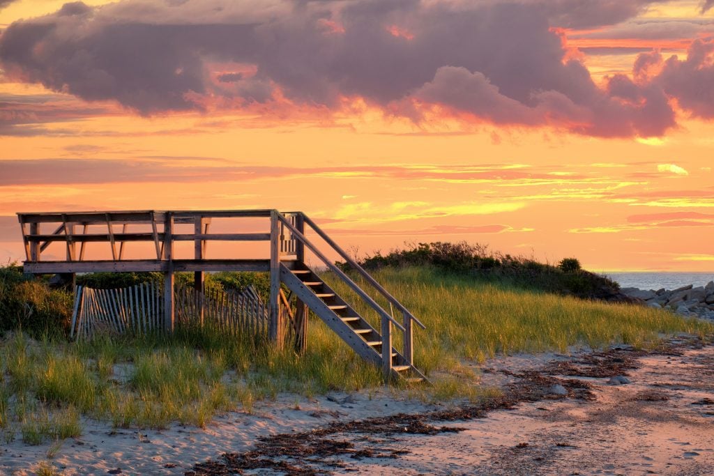 A wooden staircase and footbridge leading over the sand dunes to a beach as the sun sets, the sky colors fiery orange, pink, and purple.