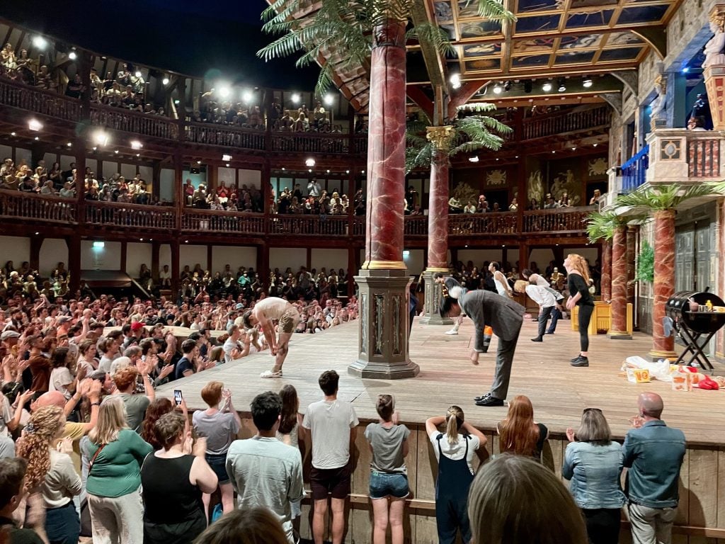 An open-air wooden theater with no roof. People standing surrounding the stage. On the stage, a cast of characters takes their bows.