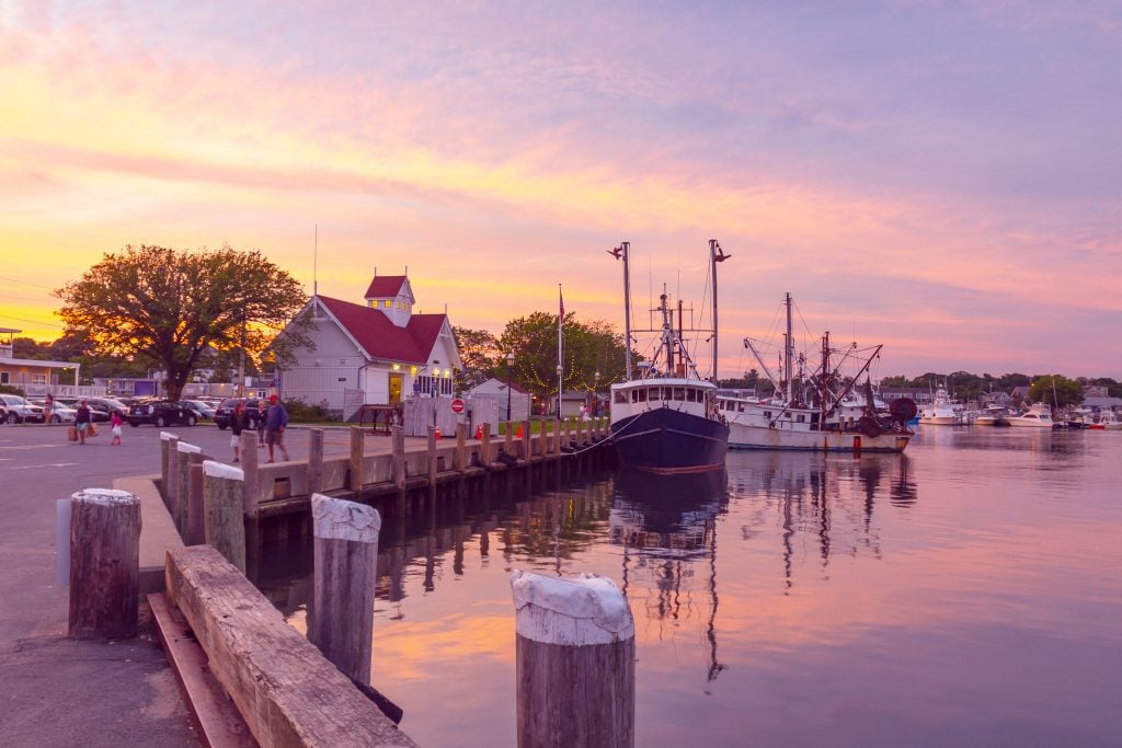 A pink sunset scene by the water in Hyannis, fishing boats parked in the harbor and people walking down the street.