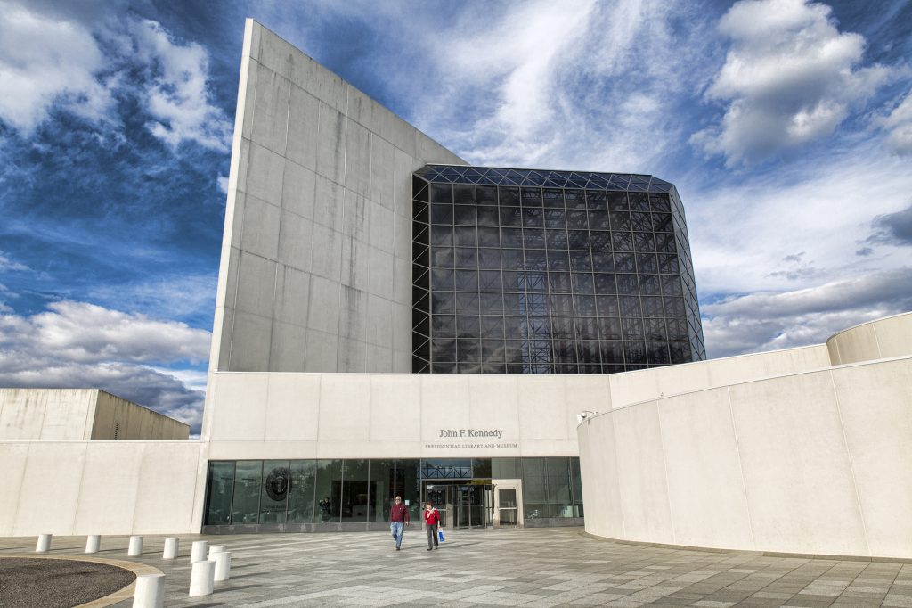 A modern white concrete building with a black glass attachment underneath a cloud-streaked blue sky.