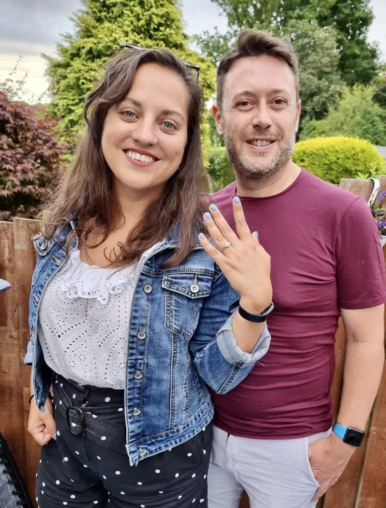 Kate and Charlie stand side by side. Kate holds up her left hand with an engagement ring on her finger, plus pale blue glittery nails. Kate wears a denim jacket, white lacy top and black shorts with white polka dots. Charlie wears a maroon t-shirt and khaki shorts.