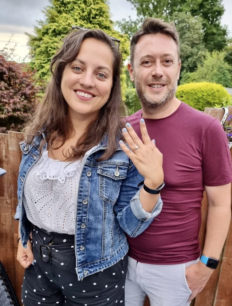 Kate and Charlie stand side by side. Kate holds up her left hand with an engagement ring on her finger, plus pale blue glittery nails. Kate wears a denim jacket, white lacy top and black shorts with white polka dots. Charlie wears a maroon t-shirt and khaki shorts.