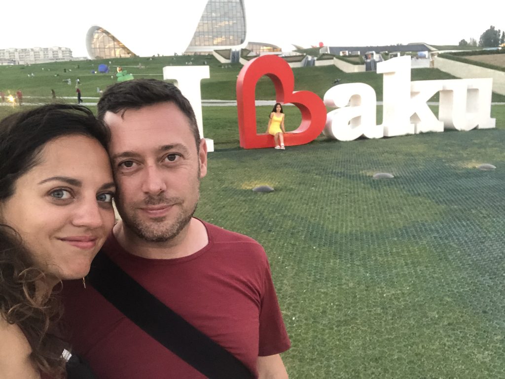 Kate and Charlie take a smiling selfie in front of big letters on grass reading I Baku. The B is a sideways red heart.