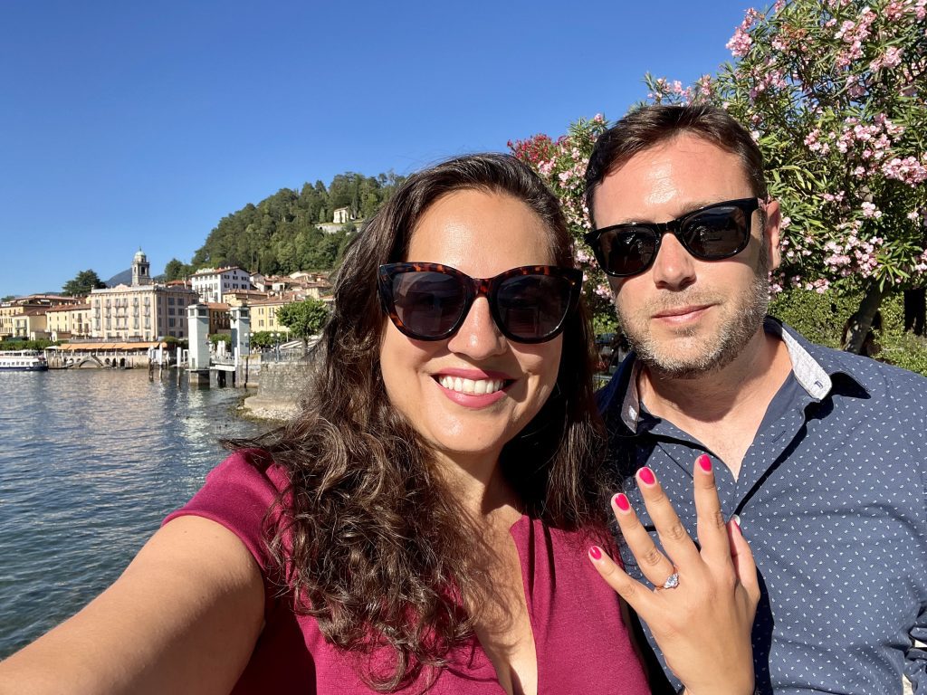 Kate and Charlie taking a smiling selfie wearing sunglasses. Kate holds up one hand and you can see an engagement ring on her finger. Behind them, Renaissance buildings set on a lake.